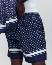 A person stands against a light gray background wearing Psycho Bunny's MENS HUNTER ALL OVER PRINT SWIM TRUNK (MB1000011), featuring a navy blue geometric and crest pattern, quick-dry fabric, and white horizontal stripes near the hems.