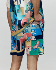 A person wears Psycho Bunny's MENS BLAISE SWIM TRUNK (MB1000028), featuring colorful tropical and beach-themed patterns—martinis, deck chairs, cactus, flowers, geometric shapes, and Bunny logos—with a matching shirt.
