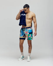 A man wears Psycho Bunny MENS BLAISE SWIM TRUNK - MB1000028, featuring colorful patterns and subtle bunny logos, paired with sneakers. He holds sunglasses and a dark towel over his shoulder, standing against a plain background.