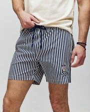 A person in Psycho Bunny MENS ZANE SWIM TRUNK - MB1000030, featuring navy and white vertical stripes and a bunny face logo on the left leg, stands against a plain background wearing a light shirt and holding a waterproof bag.