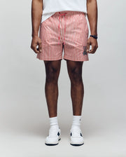 A person stands against a plain background wearing Psycho Bunny MENS ZANE SWIM TRUNK - MB1000030, a white shirt, white crew socks, and white sneakers. The shorts have red and white vertical stripes and feature a small black bunny logo. Hands relaxed at their sides.