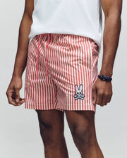 A man wears Psycho Bunny MENS ZANE SWIM TRUNK - MB1000030 featuring red and white vertical stripes, a cartoon bunny and crossbones patch, comfy no-chafe net lining, plus a white tee and dark bracelet. His hand rests casually by his side.