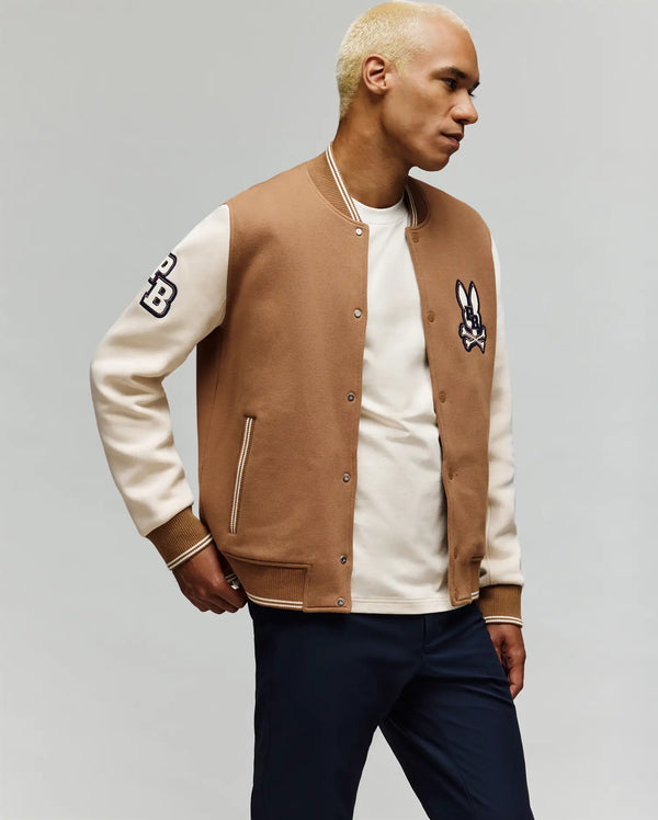 A man with bleached blond hair wears the Psycho Bunny MENS WADE KNIT BOMBER (MO1100020) in brown and cream with an embroidered rabbit patch and "PB" on the sleeve, over a white shirt and navy pants, set against a light grey background.