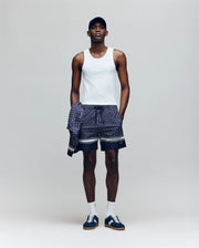 A man wears a Psycho Bunny MENS 2 PACK TANK TOP - MO1400001 in white, with patterned shorts, sneakers, white socks, a dark baseball cap, and a matching patterned shirt tied around his waist against a plain background.