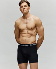 A man in Psycho Bunny MEN'S 3 PACK BOXER BRIEF - MO1400006 stands against a plain background, wearing black, premium cotton-modal boxer briefs with the Psycho Bunny logo on the waistband and a neutral expression.