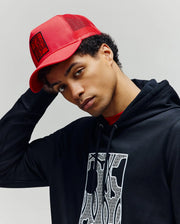 A young man wears the Psycho Bunny MENS GLENN TRUCKER CAP - MO1500004 in red and a black hoodie with a white patterned graphic, looking at the camera with a neutral expression against a plain light background.