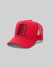 The Psycho Bunny MENS GLENN TRUCKER CAP (MO1500004) is a red mesh hat with a curved brim, adjustable snapback, and a front square patch featuring an abstract black line design, shown on a light gray background.