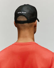A person is seen from behind against a gray background, wearing Psycho Bunny’s MENS GLENN TRUCKER CAP - MO1500004 in black mesh with branding on the back, paired with a red crewneck shirt.