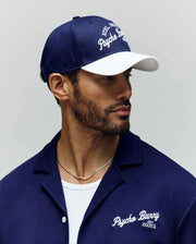 A man with a trimmed beard wears the Psycho Bunny MENS MATTEO BASEBALL CAP (MO1500008) in navy and white, paired with a matching cotton twill shirt, both featuring the embroidered Psycho Bunny wordmark. He looks to his left against a neutral background.