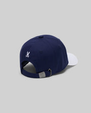 The Psycho Bunny MENS MATTEO BASEBALL CAP - MO1500008 is a navy cotton twill cap with a white brim, shown from the back. It features an adjustable strap with metal clasp and bold varsity-style embroidery above the opening on a light gray background.