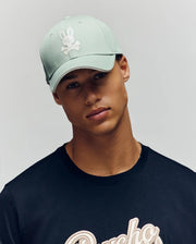A young man wears the Psycho Bunny MENS DEREK BASEBALL CAP - MO1500009 in bold light green, featuring a soft cotton build and white bunny-and-crossbones logo, paired with a black tee, against a neutral background.