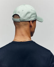 A person seen from behind wears a bold light green Psycho Bunny MENS DEREK BASEBALL CAP (MO1500009) and a gold chain necklace, with short hair and a dark shirt, set against a plain background.