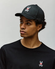 A young man wears the Psycho Bunny MENS MISCHIEF BASEBALL CAP - MO1500015 and a black shirt, both 100% cotton with the signature Mischief Bunny logo, as he looks left against a plain light gray background.
