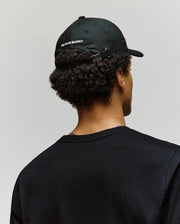 A person with curly hair, wearing a black Psycho Bunny MENS MISCHIEF BASEBALL CAP (MO1500015) made of 100% cotton and a black shirt, is shown from behind against a plain light background.