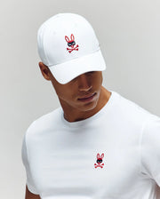 A man in a white Psycho Bunny MENS MISCHIEF BASEBALL CAP (MO1500015) and matching t-shirt, both with the red and black Mischief Bunny with crossbones design, looks downward against a plain light gray background.