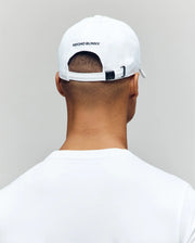 A person is shown from behind against a plain light background, wearing a white Psycho Bunny MENS MISCHIEF BASEBALL CAP (MO1500015) with "PSYCHO BUNNY" on the back, and a white 100% cotton shirt.