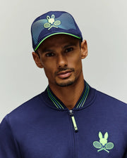 A man wears a Psycho Bunny TENNIS BALL CREW MENS CAP ENGLISH (MO1500024) in blue, with wave patterns and a tennis-themed bunny graphic, plus a matching jacket. He faces the camera against a plain light background, channeling the National Bank Open vibe.