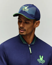 A man in a Psycho Bunny TENNIS BALL CREW MENS CAP FRENCH (MO1500025) and a matching jacket with green tennis-themed logos, including a rabbit and tennis rackets from the National Bank Open, looks downward against a light background.