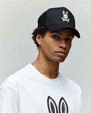 A person wears a Psycho Bunny MENS RYDER TRUCKER CAP (MO1500030) in black cotton twill with a white skull and bunny logo, paired with a white tee featuring the matching bunny logo, standing against a plain light background.
