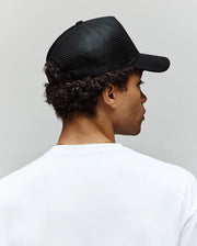 A person with short curly hair, seen from behind against a plain light background, wears a white T-shirt and a black Psycho Bunny MENS RYDER TRUCKER CAP - MO1500030 with an adjustable snapback.