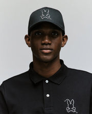 A man wears the Psycho Bunny MENS CYRUS BASEBALL CAP (MO1500045) and a black polo, both featuring the iconic white stitched bunny logo, standing against a light gray background for a blend of casual charm and modern style.