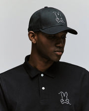 Wearing the MENS CYRUS BASEBALL CAP by Psycho Bunny and a matching black polo, each with the iconic white embroidered bunny logo, a person channels classic style while looking down against a plain background.
