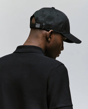 A man wearing the Psycho Bunny MENS CYRUS BASEBALL CAP (MO1500045) and a black collared shirt is seen from behind against a plain light background, giving the scene a casual, nautical vibe.