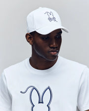 A man wears the Psycho Bunny MENS CYRUS BASEBALL CAP (MO1500045) and a matching T-shirt, both with the brand’s iconic outlined bunny and crossed bones, looking down neutrally against a plain background.