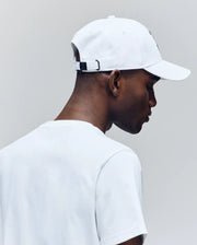 A person is seen from behind and in profile wearing the Psycho Bunny MENS CYRUS BASEBALL CAP (MO1500045) and a white t-shirt, looking down against a light grey background—evoking effortless coastal charm.