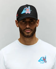 A bearded man stands against a light gray background wearing a Psycho Bunny MENS MONTREAL BASEBALL CAP - MO1500048 in black and a white 100% cotton t-shirt, both featuring an embroidered Bunny with blue triangle and pink accents.