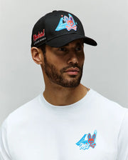 A man with a short beard wears a Psycho Bunny 100% cotton white shirt and the MENS MONTREAL BASEBALL CAP - MO1500048, both featuring colorful cartoon bunny graphics and blue geometric designs. The black cap has red embroidery on the side. Background is gray.
