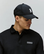 A man wearing a black collared shirt and the Psycho Bunny MENS FORREST SPORT CAP (MO1500051), featuring an adjustable strap and a white bunny-and-crossbones logo, looks down to the side against a plain background.