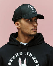 A man stands in front of a pink background, wearing the Psycho Bunny MENS ARCHIVE BASEBALL CAP (MO1500002) and a black 100% cotton hoodie featuring an embroidered bunny logo and "Psycho Bunny" text, looking to the side.