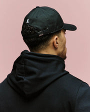 A person wearing a black 100% cotton hoodie and the Psycho Bunny MENS ARCHIVE BASEBALL CAP (MO1500002) is shown from behind against a plain pink background.