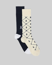 The Psycho Bunny MENS 2 PACK DRESS SOCK (MO2200005) includes one black crew sock with a white cartoon skull and bones, and one cream sock with a diamond pattern, skull motifs, and "P E" in black; both have white heels and toes.