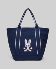 The Psycho Bunny MENS CANVAS TOTE BAG - MO2400016 is a navy blue tote with white handles and stripes, plus an embroidered skull and crossbones with pink bunny ears. Made for durability, it’s a stylish match for premium athleisure. Light gray background.