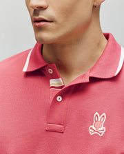 A man wears a red Psycho Bunny Mens Fenley Pique Polo (MT0100009) made from soft Pima cotton, with white trim and an embroidered Bunny and crossbones chest logo. Only his lower face and upper torso are visible.