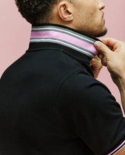 A person adjusts the high collar of Psycho Bunny's MENS ARCHIVE CLASSIC BUNNY PIQUE POLO (MT0100010), a black shirt with pink, white, and black stripes and an intarsia-knit undercollar, against a pink background. Only their lower face and upper torso are visible.