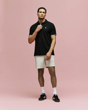 A man stands against a pink background wearing Psycho Bunny’s MENS ARCHIVE CLASSIC BUNNY PIQUE POLO - MT0100010, light beige shorts, white socks, and black Adidas sneakers. He holds the polo’s collar with one hand and looks forward.