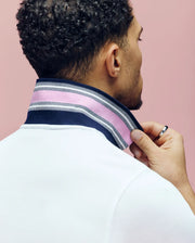 A man with curly hair, seen from behind, lifts the collar of his white Psycho Bunny MENS ARCHIVE CLASSIC BUNNY PIQUE POLO to reveal a striped navy, pink, and white pattern. He wears a silver ring and stands against a pink background.