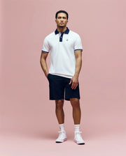 A man stands against a pink background wearing the Psycho Bunny MENS ARCHIVE CLASSIC BUNNY PIQUE POLO - MT0100010, featuring white Pima cotton with a dark collar, paired with black shorts, white socks, and high-top sneakers.