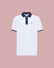 The Psycho Bunny MENS ARCHIVE CLASSIC BUNNY PIQUE POLO (MT0100010) is a white Pima cotton short-sleeve polo with a navy collar, button placket, sleeve trim, and an embroidered Bunny logo on the chest, shown on a pink background.