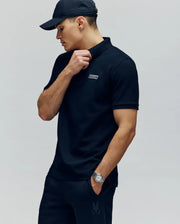 A man in a Psycho Bunny MENS AXTON PIQUE POLO (MT0100011), black cap, and pants stands against a plain background, wearing a watch on his left wrist and touching his collar with his right hand while looking to the side.