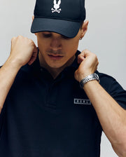 A person in a black Psycho Bunny MENS AXTON PIQUE POLO—featuring the iconic skull and bunny ears logo—wears a black cap, silver watch, adjusts their collar, and looks down.