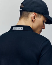 A person is seen from behind, slightly turned, wearing a black Psycho Bunny MENS AXTON PIQUE POLO - MT0100011 and a black cap, with the white "Psycho Bunny" label visible on the back of the collar.