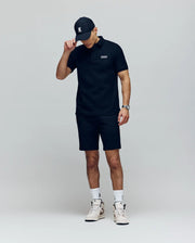 A man in a Psycho Bunny MENS AXTON PIQUE POLO (MT0100011), black shorts, a black cap, and white high-top sneakers stands against a plain light background, tilting his cap downward with one hand.