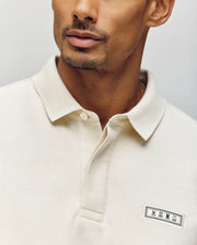 A man wears the Psycho Bunny MENS AXTON PIQUE POLO (MT0100011) in cream Pima cotton, featuring a concealed button placket and a jacquard logo patch with three minimalistic animal icons. The image shows only his lower face and upper torso.