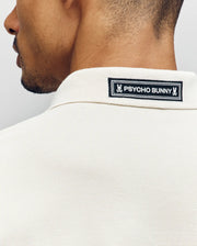 Close-up of the back of a man's neck and shoulders wearing the Psycho Bunny MENS AXTON PIQUE POLO (MT0100011) in cream, featuring a black-and-white jacquard-woven logo patch sewn onto the collar.