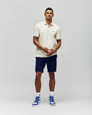 A man stands against a plain background wearing the Psycho Bunny MENS AXTON PIQUE POLO - MT0100011 in cream, paired with navy shorts, white socks, and white and blue sneakers. He faces forward with his hands clasped in front of him.