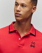 A young man wears the Psycho Bunny MENS JUDE PIQUE POLO - MT0100012 in bright red, made from soft Pima cotton with an embroidered Bunny skull and crossbones logo on the chest, gazing slightly to the side against a plain backdrop.